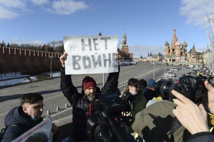 A man holds a poster which reads \"No war\" as people lay flowers near the place where Russian opposition leader Boris Nemtsov was gunned down, with the Kremlin Wall, left, the Spaskaya Tower, center, and St. Basil\'s in the background, in Moscow, Russia, Sunday, Feb. 27, 2022. (AP Photo/Denis Kaminev)